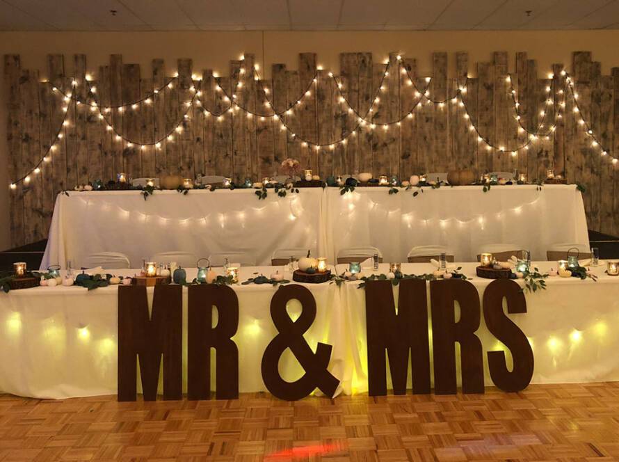 Rustic wedding head table at The Pavilion with string lights, wood backdrop, and large MR & MRS letters.