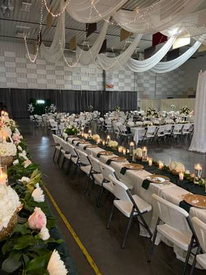 Wedding reception setup in The Pavilion Expo Hall with long banquet tables, floral arrangements, candles, and draped ceiling lights.