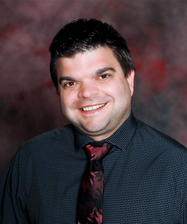 Professional headshot of The Pavilion Administrative Assistant, Jeff Keasler-Bird, wearing a dark shirt and patterned tie against a studio backdrop.