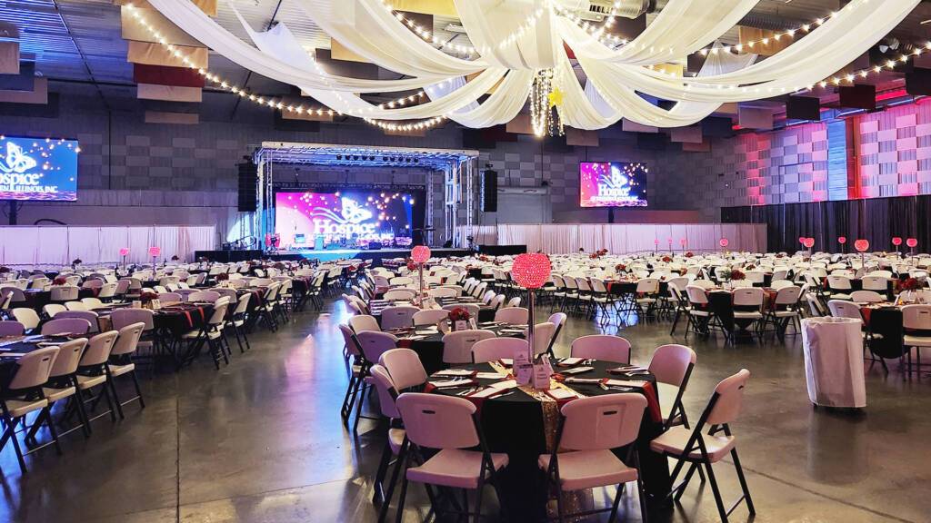 The Pavilion Expo Hall decorated for a large banquet event space rental with round tables, stage, hanging drapery, and colorful lighting.